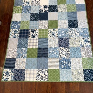 May include: A patchwork quilt featuring a variety of blue, green, and white patterned squares. The quilt has a green border and is made of various fabrics, including floral, striped, and dotted designs. The quilt is displayed on a wooden floor.