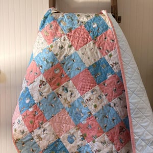May include: A patchwork quilt with a light blue backing and a pink gingham border. The quilt's top features squares of pink, light blue, and white fabric, each with a different illustration of rabbits and other characters.