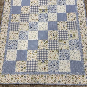 May include: A patchwork quilt with a blue and white color scheme. The quilt features squares of gingham, polka dots, and floral patterns. The border has a repeating pattern of small animals and plants.