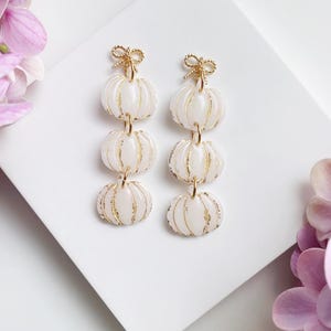 May include: A pair of white and gold pumpkin-shaped earrings. Each earring features three stacked pumpkins with gold accents and a gold bow at the top. The earrings are displayed on a white surface.