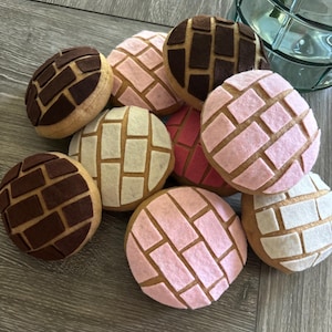 May include: A collection of colorful, plush concha bread. The soft, round pastries feature a variety of colors, including brown, pink, and white, with a textured, patterned topping. These decorative items are perfect for a bakery or kitchen theme.