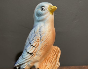 May include: A blue and brown ceramic bird figurine perched on a branch with a pink flower. The bird has a yellow beak and black eyes.