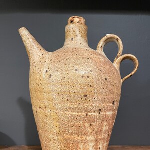 May include: A brown and white ceramic pitcher with a cork stopper and two handles. The pitcher has a speckled glaze and a rustic look.