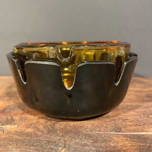 May include: A black and amber glass ashtray with a unique, wavy design. The ashtray has a wide, shallow bowl and a flared rim.