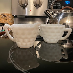 May include: Two white milk glass creamer pitchers with a textured design. The pitchers are on a black electric stovetop.