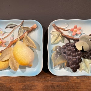 May include: Two decorative ceramic wall plaques with 3D fruit designs. One plaque features a branch of lemons with green leaves and pink flowers. The other plaque features a branch of grapes with green leaves and a pink flower.
