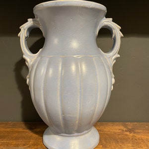 May include: A light blue ceramic vase with two handles and a ribbed design. The vase has a slightly iridescent finish.