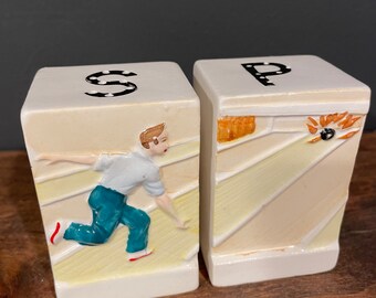 May include: A set of two white ceramic salt and pepper shakers shaped like bowling pins. The shakers are decorated with a bowling scene, including a bowler, bowling pins, and a bowling ball. The shakers are labeled "S" and "P" for salt and pepper.