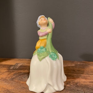 May include: A porcelain bell with a white tulip-shaped base. A small boy figurine sits on top of the bell, holding a green stem with shamrock leaves.