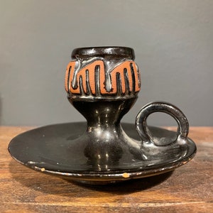 May include: Black ceramic candle holder with a brown geometric design and a handle. The holder has a small, round base.