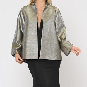 Puede incluir: Chaqueta metalizada dorada y plateada con frente abierto y cuello alto. La chaqueta tiene mangas largas y un aspecto texturizado. La chaqueta se lleva sobre un vestido negro.