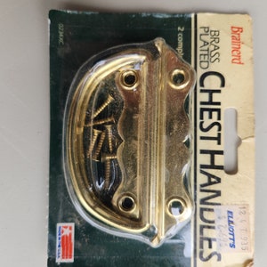 Könnte beinhalten: Eine Packung mit zwei vermessingten Truhengriffen mit Schrauben. Die Verpackung ist grün und weiß mit dem Markennamen "Brainard" und dem Text "Vermessingte Truhengriffe".