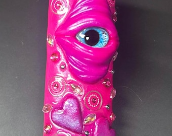 May include: A cylindrical lighter case in vibrant hot pink, featuring a sculpted three-dimensional eye with a realistic blue iris and black pupil. The surface is decorated with small purple heart shapes and numerous clear, reflective rhinestones.