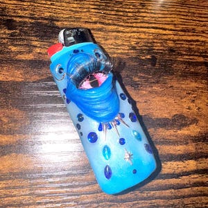 May include: A blue Bic lighter decorated with a sculpted blue mouth featuring long black eyelashes and a pink interior. The lighter is embellished with various blue and teal rhinestones, small silver spikes, and a silver star-shaped charm.
