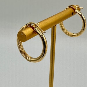 May include: A pair of gold hoop earrings with a textured design. The earrings are displayed on a gold stand.