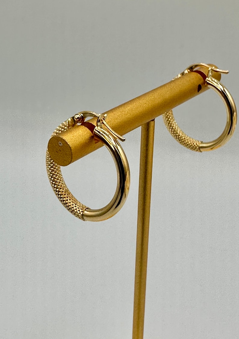 May include: A pair of gold hoop earrings with a textured design. The earrings are displayed on a gold stand.