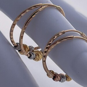 May include: Gold hoop earrings with a textured design. Each earring features a series of small, decorative beads in gold, silver, and rose gold. The earrings are displayed on a white surface.