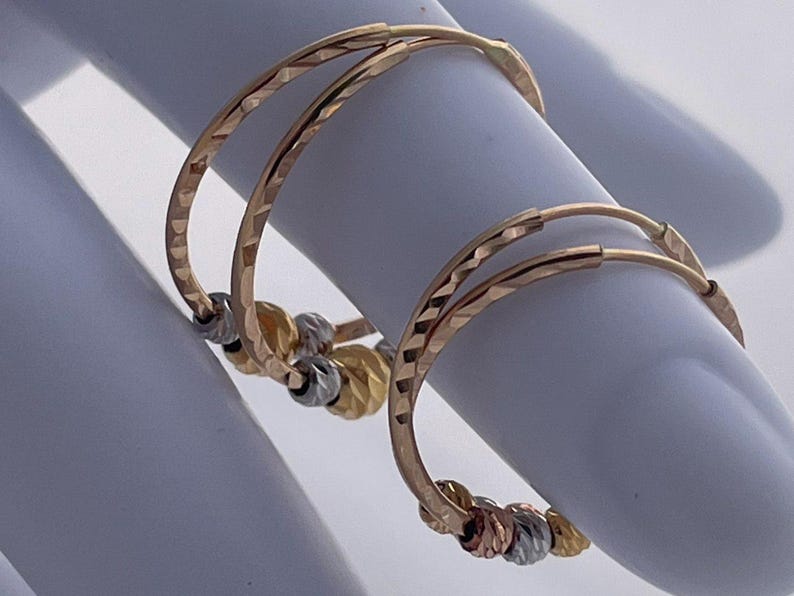 May include: Gold hoop earrings with a textured design. Each earring features a series of small, decorative beads in gold, silver, and rose gold. The earrings are displayed on a white surface.