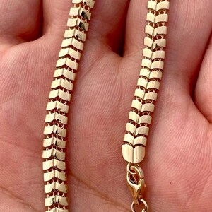 May include: A gold-colored chain necklace with a unique, textured design. The chain is made of small, rectangular links arranged in a repeating pattern. The necklace is shown against a skin-toned background and has a clasp.