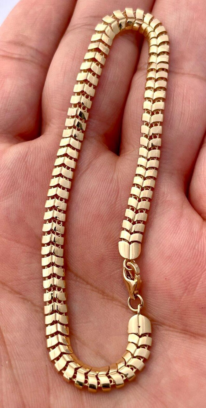 May include: A gold-colored chain necklace with a unique, textured design. The chain is made of small, rectangular links arranged in a repeating pattern. The necklace is shown against a skin-toned background and has a clasp.