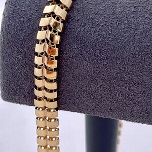 May include: A gold-colored bracelet with a unique chevron design. The bracelet is made of rectangular links that are connected to form a flexible chain. The bracelet is displayed on a gray velvet surface.