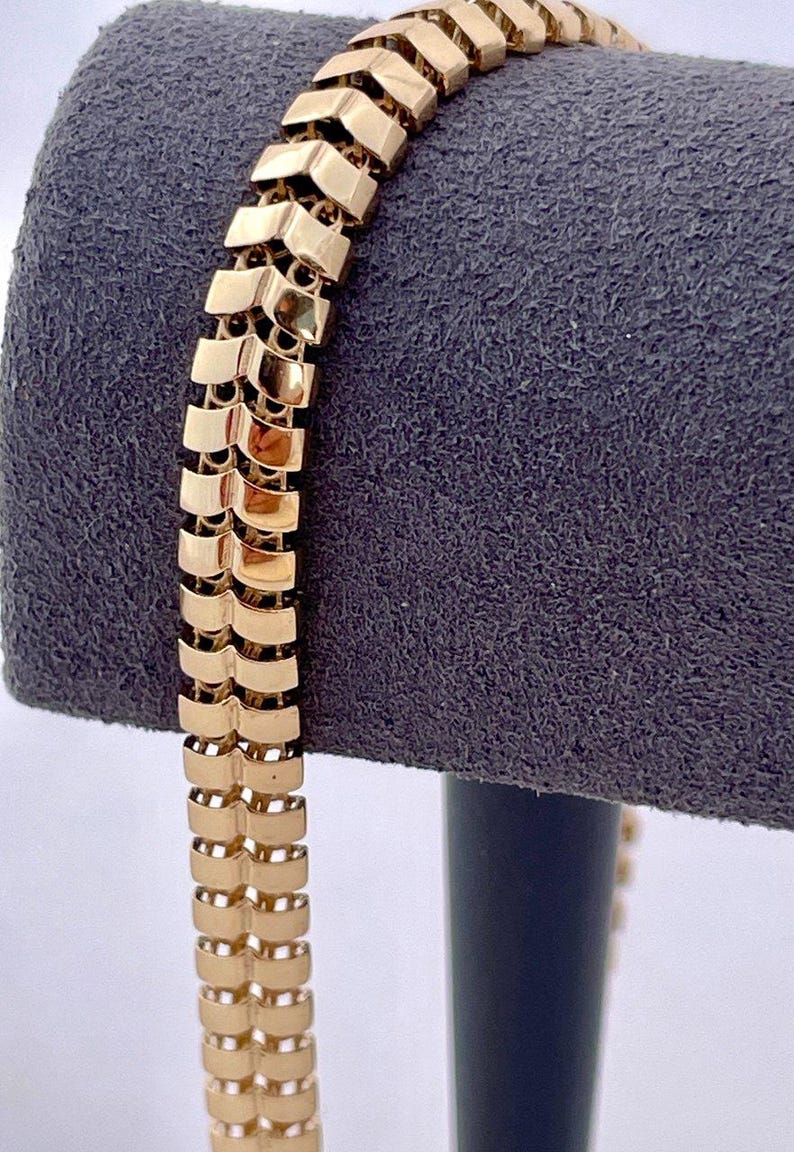 May include: A gold-colored bracelet with a unique chevron design. The bracelet is made of rectangular links that are connected to form a flexible chain. The bracelet is displayed on a gray velvet surface.