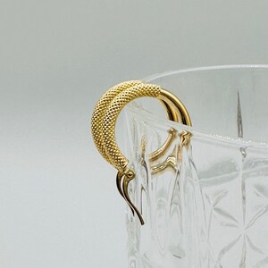 May include: A pair of gold hoop earrings with a textured surface. The earrings are hanging from a clear glass container.