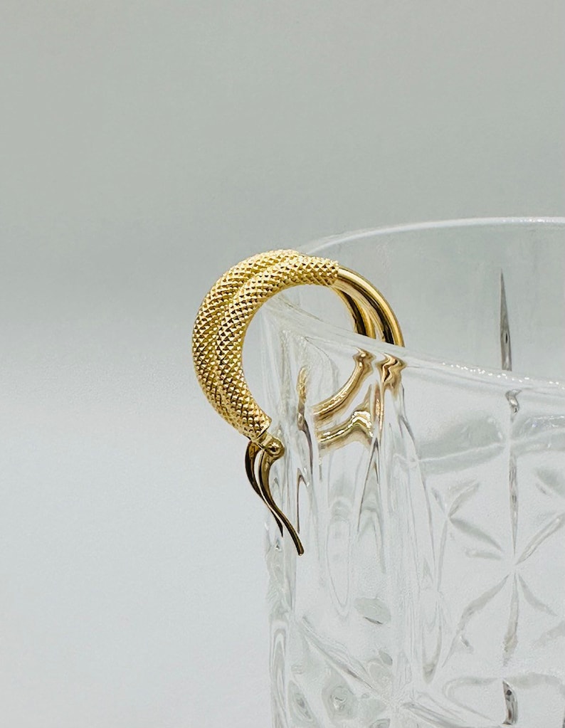May include: A pair of gold hoop earrings with a textured surface. The earrings are hanging from a clear glass container.