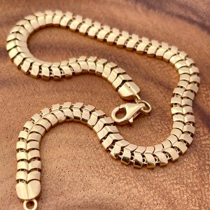 May include: A gold-colored bracelet with a unique, textured design. The bracelet features a series of interconnected, rectangular links, creating a flexible and eye-catching pattern. The clasp is a lobster claw style. The jewelry is displayed on a wooden surface.