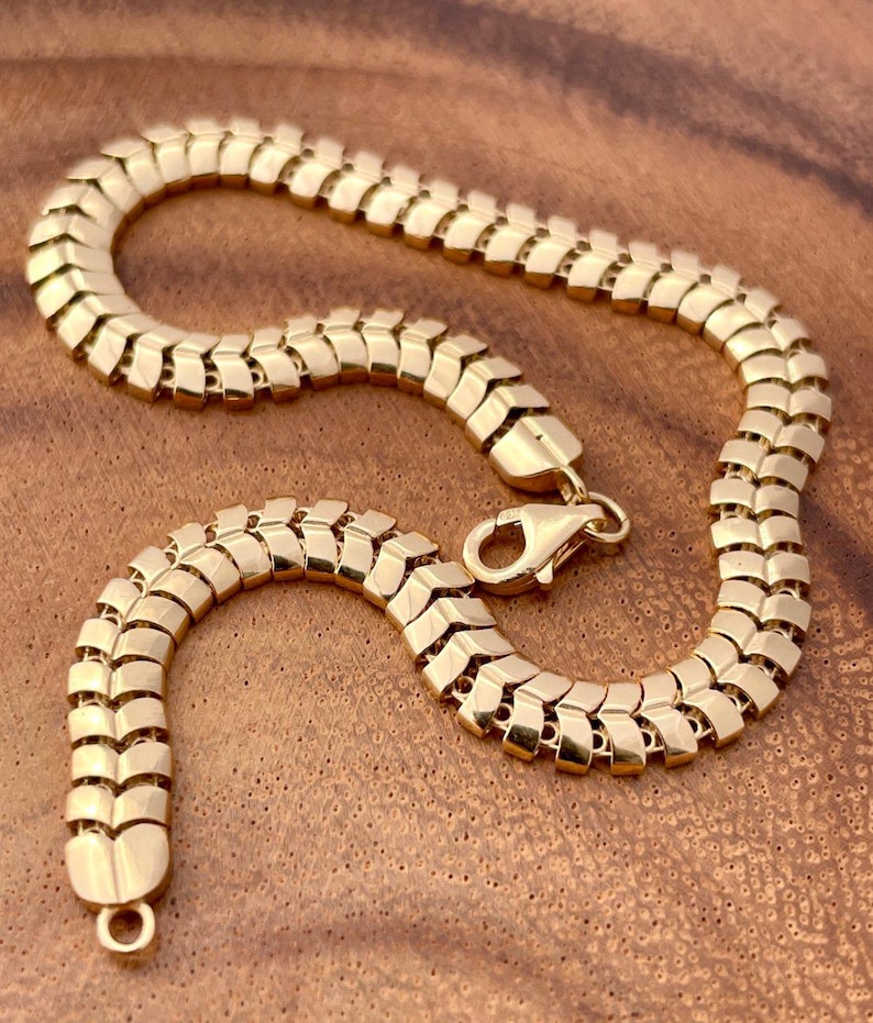 May include: A gold-colored bracelet with a unique, textured design. The bracelet features a series of interconnected, rectangular links, creating a flexible and eye-catching pattern. The clasp is a lobster claw style. The jewelry is displayed on a wooden surface.