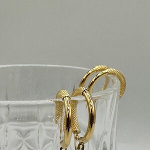 May include: A pair of gold hoop earrings with a textured finish. The earrings are hanging from the edge of a clear glass container.