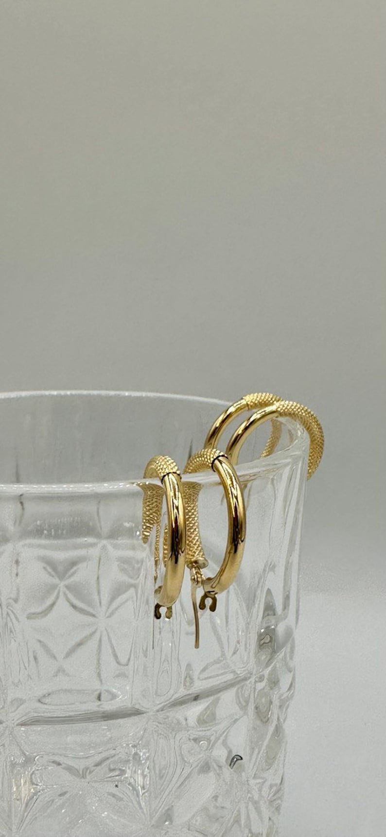 May include: A pair of gold hoop earrings with a textured finish. The earrings are hanging from the edge of a clear glass container.
