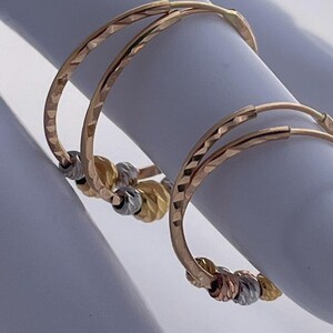 May include: A pair of gold hoop earrings with a textured surface. Each earring features a series of small, decorative beads in gold, silver, and rose gold. The earrings are displayed on a white surface.
