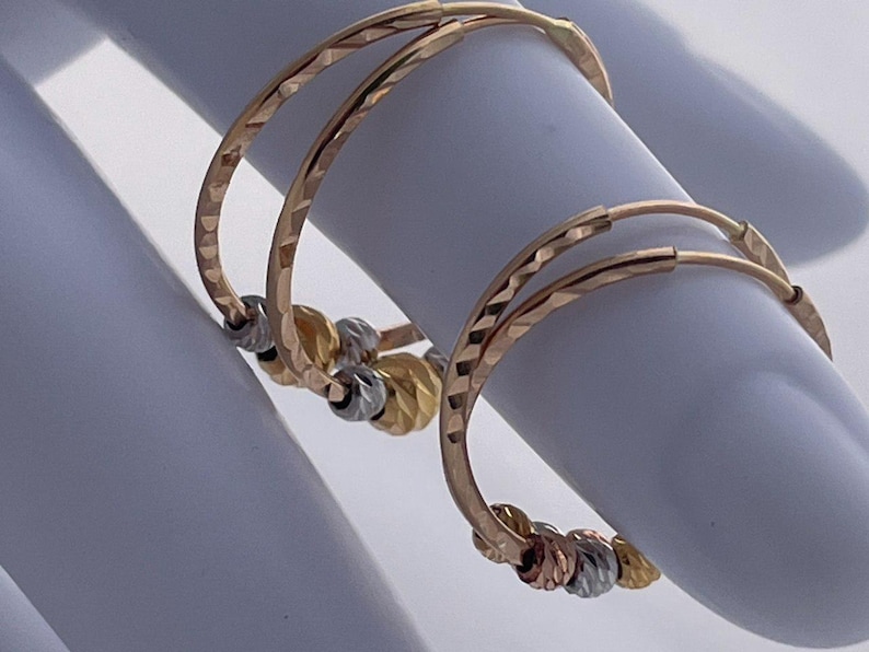 May include: A pair of gold hoop earrings with a textured surface. Each earring features a series of small, decorative beads in gold, silver, and rose gold. The earrings are displayed on a white surface.