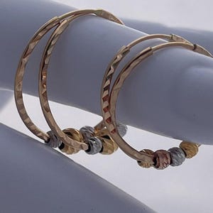 May include: A pair of gold hoop earrings with a textured design. Each earring features a series of small, decorative beads in gold, silver, and rose gold. The earrings are displayed on a light blue surface.