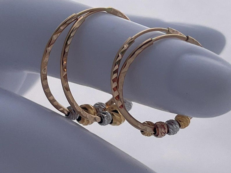 May include: A pair of gold hoop earrings with a textured design. Each earring features a series of small, decorative beads in gold, silver, and rose gold. The earrings are displayed on a light blue surface.