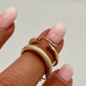 May include: A pair of gold hoop earrings with a textured surface. The earrings are shown on a finger.