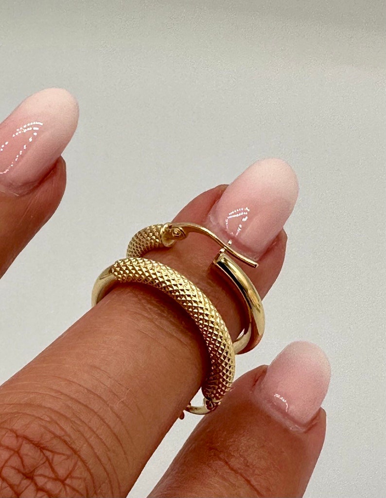 May include: A pair of gold hoop earrings with a textured surface. The earrings are shown on a finger.
