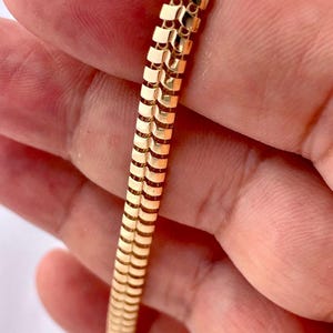 May include: A gold-colored bracelet with a textured, chevron-like pattern. The bracelet is made of small, rectangular links that are connected to form a flexible band. The bracelet is held in a person's hand.