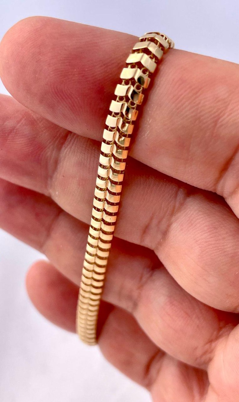 May include: A gold-colored bracelet with a textured, chevron-like pattern. The bracelet is made of small, rectangular links that are connected to form a flexible band. The bracelet is held in a person's hand.