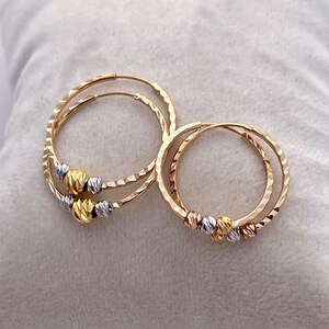 May include: Two pairs of gold hoop earrings. Each earring has a textured, hammered finish and is adorned with a cluster of three small, textured beads in gold, silver, and a third color. The earrings are displayed on a soft, light-colored surface.