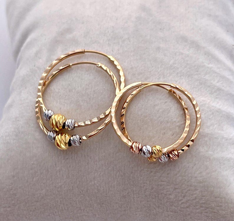 May include: Two pairs of gold hoop earrings. Each earring has a textured, hammered finish and is adorned with a cluster of three small, textured beads in gold, silver, and a third color. The earrings are displayed on a soft, light-colored surface.