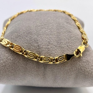 May include: A gold bracelet featuring an S-shaped link pattern. The bracelet is presented on a grey surface. The clasp is visible, and the piece appears to be made of metal.