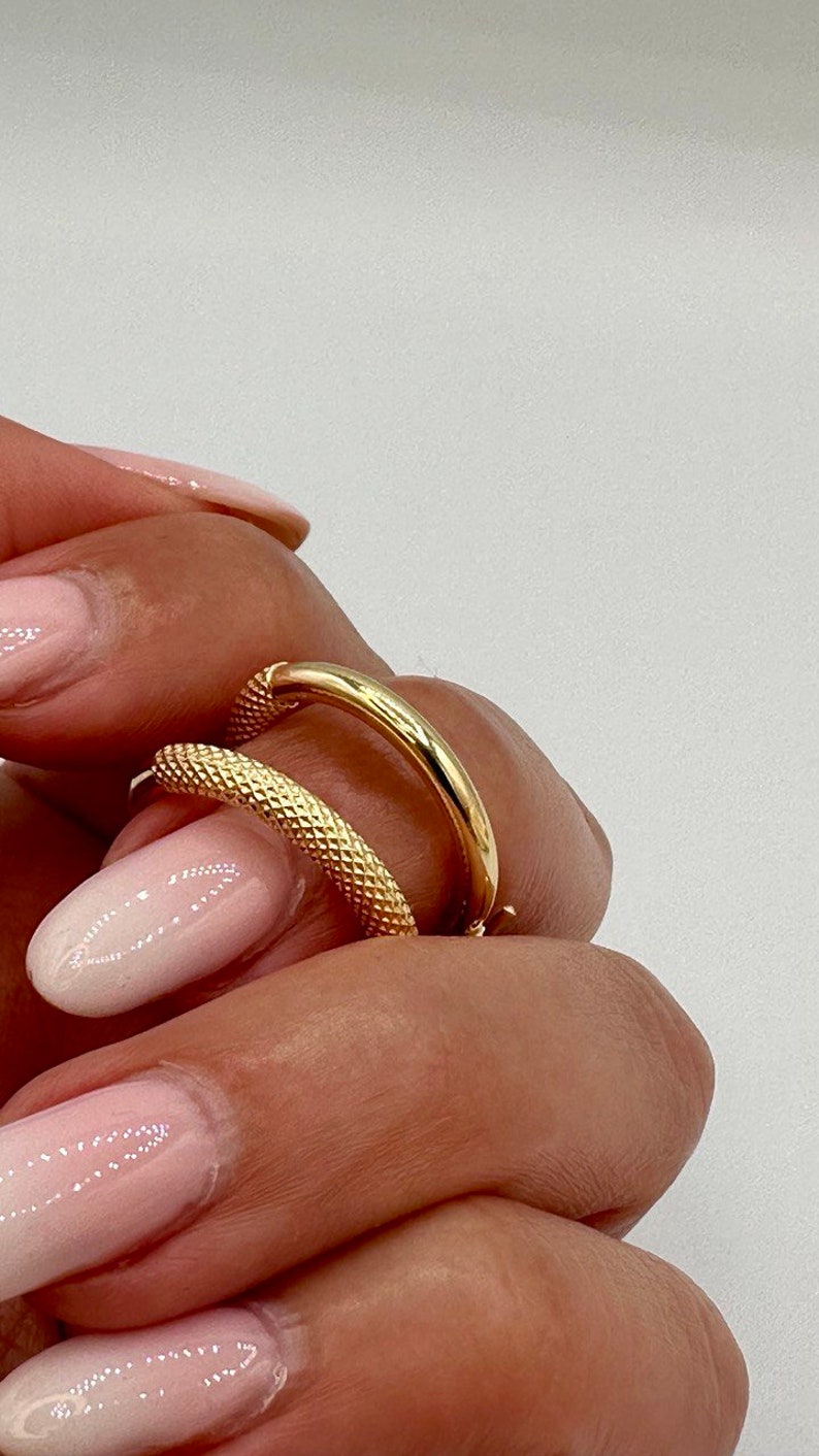 May include: Two gold hoop earrings, one with a smooth finish and the other with a textured finish. The earrings are shown on a hand with pink nail polish.