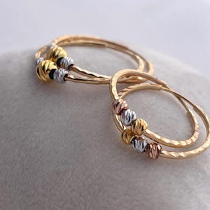 May include: Gold hoop earrings with textured bands and decorative beads in gold, silver, and rose gold tones. The earrings are displayed on a soft, white surface, showcasing their intricate design and color variations.