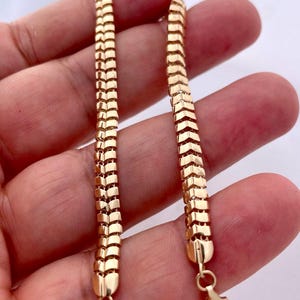 May include: A gold-colored chain bracelet with a unique, textured design. The bracelet features a series of rectangular links arranged in a chevron pattern. The bracelet is held in a hand, showcasing the intricate details and the warm, golden hue of the metal.
