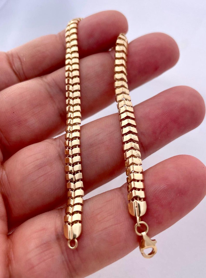 May include: A gold-colored chain bracelet with a unique, textured design. The bracelet features a series of rectangular links arranged in a chevron pattern. The bracelet is held in a hand, showcasing the intricate details and the warm, golden hue of the metal.