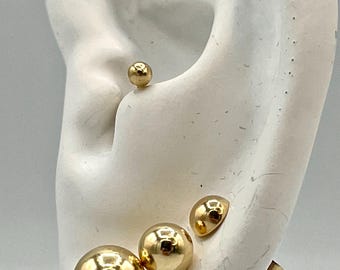 Può includere: Un set di quattro orecchini a lobo sferici color oro di varie dimensioni, esposti su un modello di orecchio bianco. Gli orecchini presentano una finitura metallica liscia e lucida con una superficie riflettente, inclusa una piccola sfera in alto e tre più grandi.