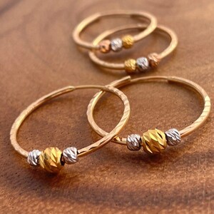 May include: Gold hoop earrings with textured detailing and three decorative beads. The beads are gold and silver in color. The earrings are displayed on a brown wooden surface.