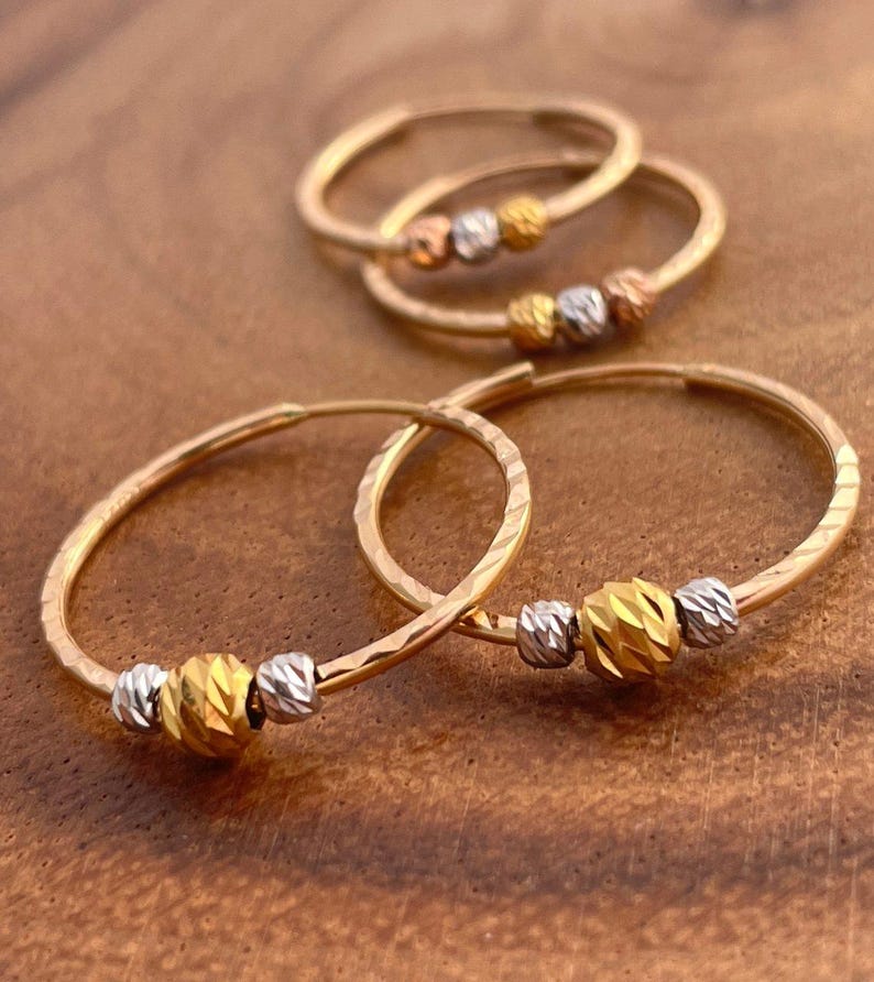 May include: Gold hoop earrings with textured detailing and three decorative beads. The beads are gold and silver in color. The earrings are displayed on a brown wooden surface.